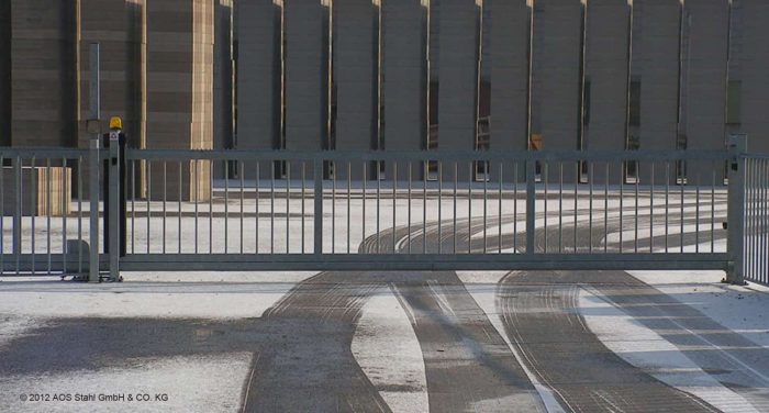 En lukket Skydeport "EasyStar" metalsikkerhedslåge med lodret stang eller panelhegn står foran en snedækket jord og viser dækspor, der fører igennem. Baggrunden viser en bygning med høje, lodrette arkitektoniske elementer, der skaber en kold og vinterlig scene. © 2012 AOS Stahl GmbH & CO. KG.