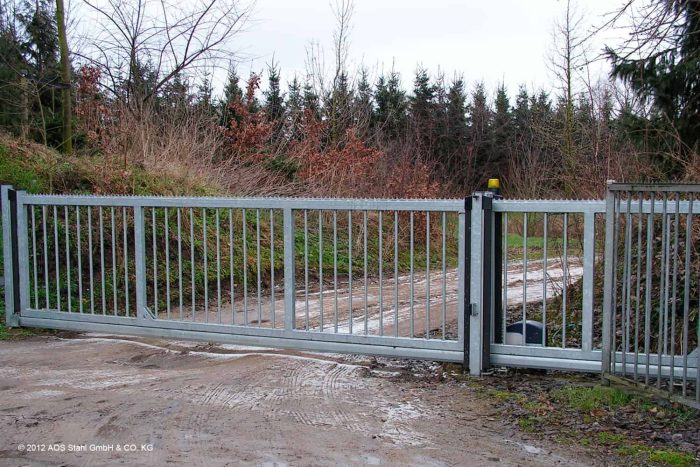 En skydeport "EasyStar"-skydeport i metal, lukket og dækket af mudder og dækspor, blokerer en snavset indkørsel omgivet af tilgroet græs og træer. Tæt løv og en række af stedsegrønne træer giver en følelse af både industri og privat afsondrethed i baggrunden.