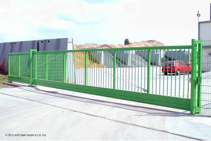 En Skydeport "EasyStar"-skydesikkerhedslåge med lodrette stænger og fås i længder fra 3 til 9,5 meter er installeret ved indgangen til et industrielt anlæg. Den grønne port løber på et spor og har en betonbelagt grund under sig. I baggrunden ses bunker af materialer og industribygninger.