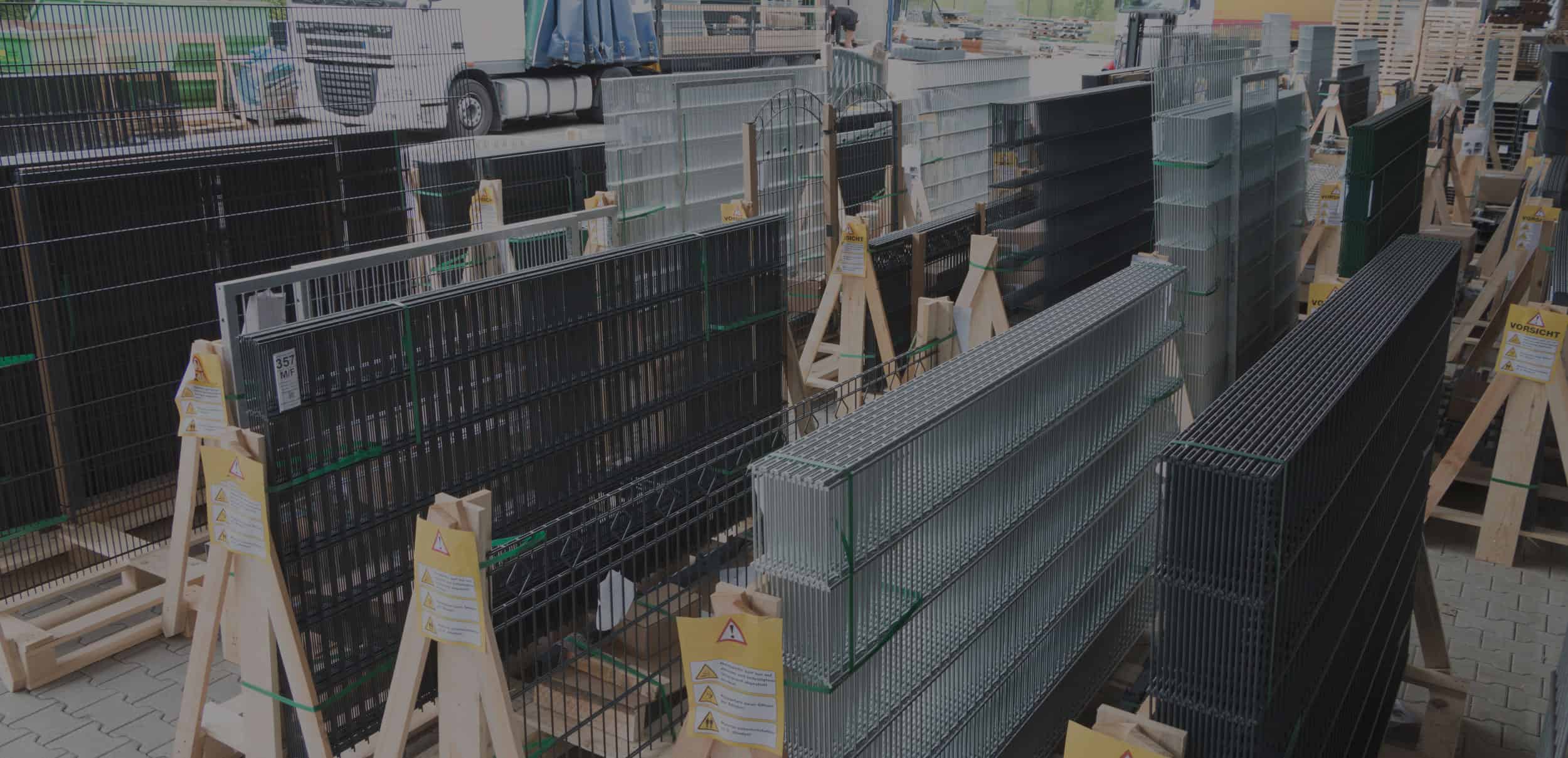 Stabler af forskellige metal hegnspaneler vises oprejst på træstandere i et udendørs opbevaringsområde. Adskillige typer hegn, der bare venter på, at løftet er på plads, er organiseret side om side med forskellige størrelser og farver lige fra sort til sølv. En lastbil er parkeret i baggrunden.