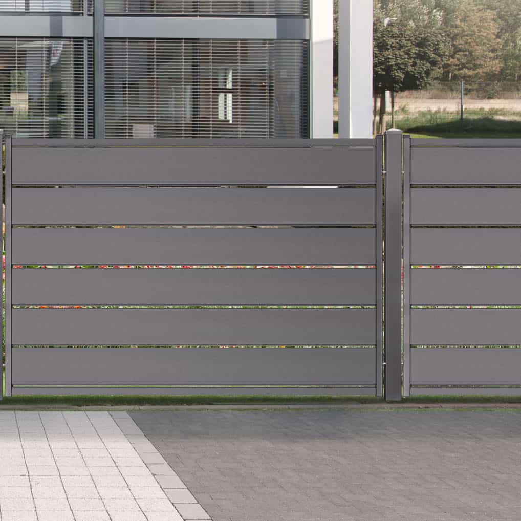 Et moderne, slankt design Hegn Pulverlakeret Aluminium Antracit Komplet - Type 1 står foran en moderne bygning med store vinduer og en græsplæne med nogle træer i baggrunden. Forgrunden har flisebelægning.