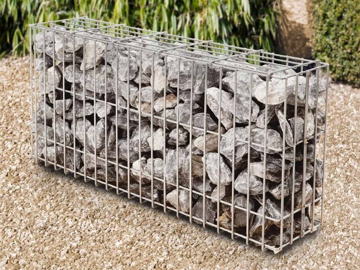 En gabionhegn, nærmere bestemt Panelhegn "FUNDA" 2D OVERFL.Behandl?, fyldt med store, uregelmæssigt formede grå sten ligger på en grusbelagt grund, med grønne buske og planter i baggrunden.