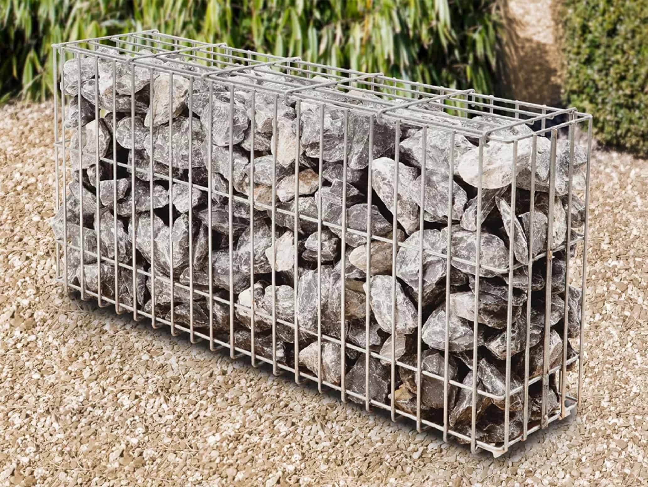 En gabionhegn, nærmere bestemt Panelhegn "FUNDA" 2D OVERFL.Behandl?, fyldt med store, uregelmæssigt formede grå sten ligger på en grusbelagt grund, med grønne buske og planter i baggrunden.