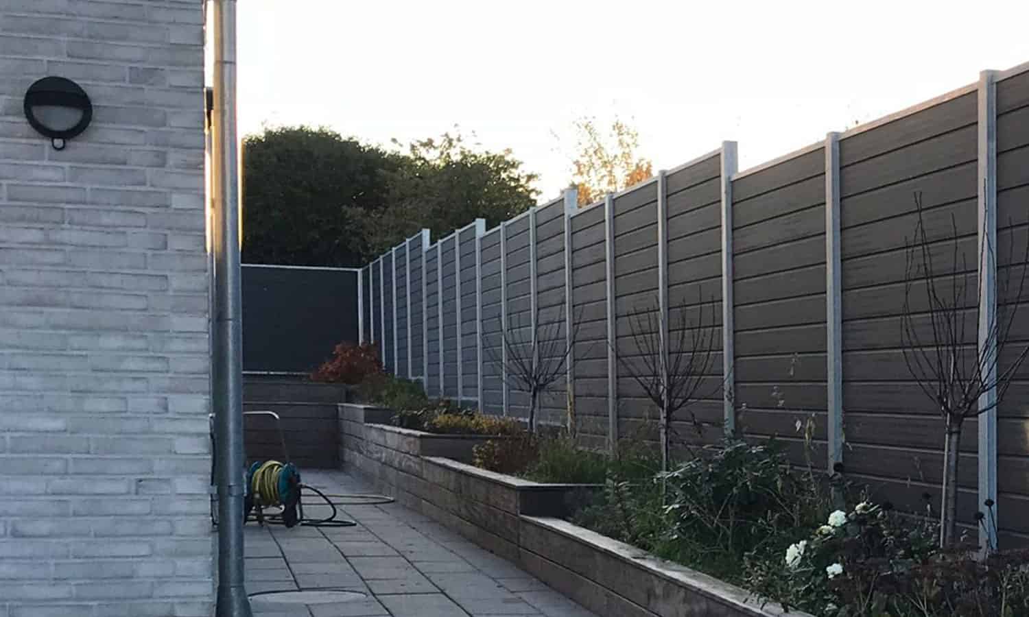 En smal brolagt havegang mellem en hvid murstensbygning til venstre og en høj, grå hegn til højre. Langs Hegn er hævede blomsterbede med unge træer og planter. En pænt oprullet grøn haveslange er sat op på jorden nær bygningen.