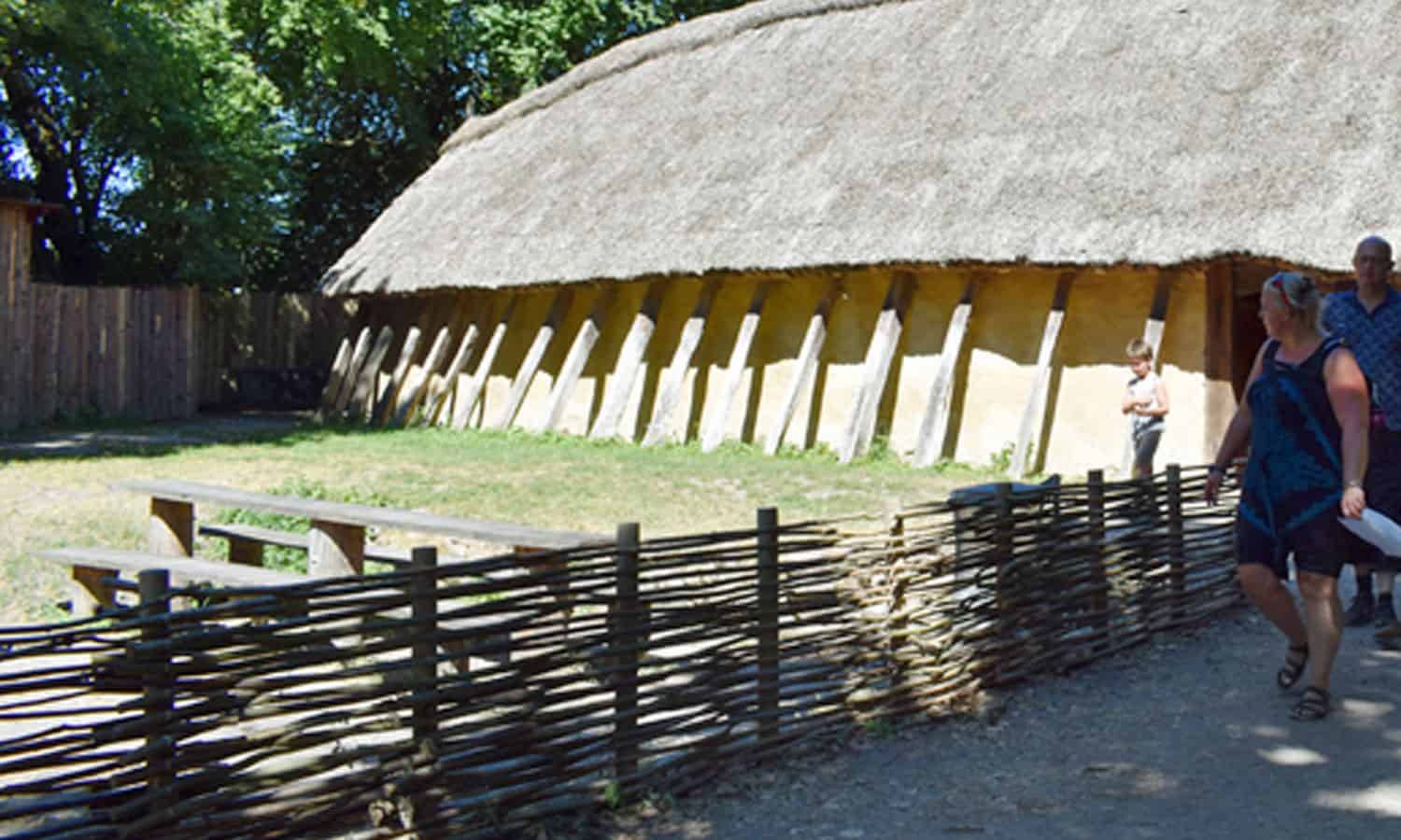 En gruppe mennesker går forbi en lang bygning med stråtag med træbjælker og skrånende tag. I forgrunden ses en vævet træhegn. Træer og grønt omgiver landskabet på en solskinsdag.