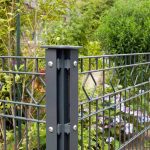 Et nærbillede af et sort metalhavehegn med geometriske diamantmønstre lavet af Stålstolper Hjørne 60x40mm med dækliste TYPE A Komplet (Ideel til nedstøbning). Hegnet er omgivet af frodigt grønt løv, inklusive forskellige planter og små blomster, hvilket skaber en rolig havestemning i baggrunden.