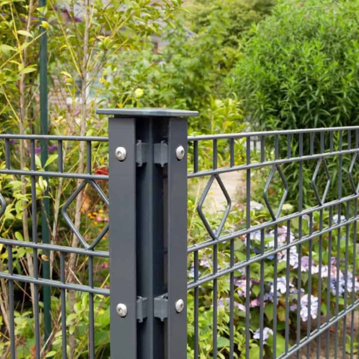 Et nærbillede af et sort metalhavehegn med geometriske diamantmønstre lavet af Stålstolper Hjørne 60x40mm med dækliste TYPE A Komplet (Ideel til nedstøbning). Hegnet er omgivet af frodigt grønt løv, inklusive forskellige planter og små blomster, hvilket skaber en rolig havestemning i baggrunden.