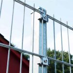 Nærbillede af et galvaniseret metalhegn med lodrette og vandrette stænger, fastgjort med bolte til en central, vandret stolpe. "Holdeplade med hul" lavet af rustfrit stål har præcise hulplaceringer. I baggrunden er der en delvis udsigt over en rød bygning og grønne træer mod en klar blå himmel.