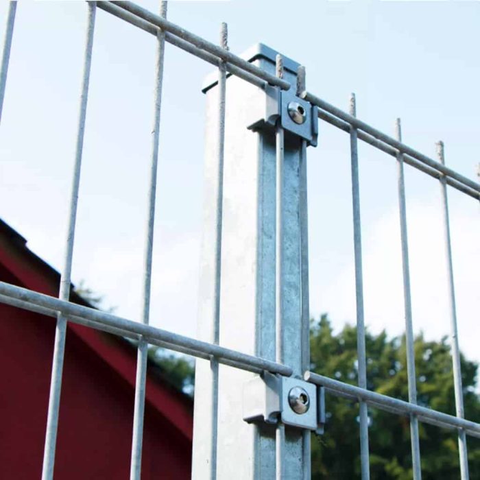 Nærbillede af et galvaniseret metalhegn med lodrette og vandrette stænger, fastgjort med bolte til en central, vandret stolpe. "Holdeplade med hul" lavet af rustfrit stål har præcise hulplaceringer. I baggrunden er der en delvis udsigt over en rød bygning og grønne træer mod en klar blå himmel.