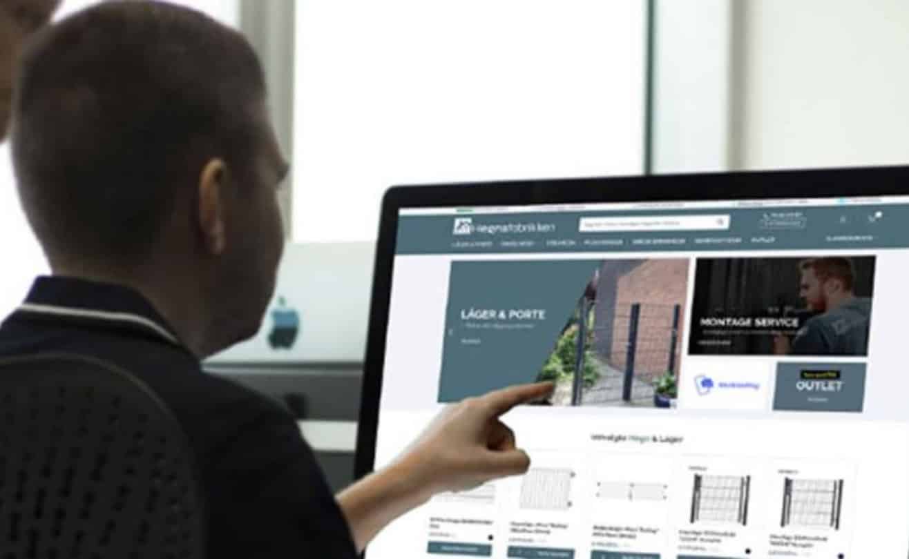 En person sidder og peger på en computerskærm, der viser en hjemmeside med speciale i hegn og porte. Hjemmesiden, hegn online, indeholder navigationslinks, billeder og bannere om forskellige produkter og tjenester for at hjælpe dig med at spare tid og penge.
