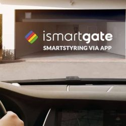 Udsigt inde fra en bil, der nærmer sig en åben garage med "ismartgate" og "SMARTSTYRING VIA APP" skrevet på indervæggen. Billedet, med en skyder, understreger brugen af en smart enhed til at styre garageporten, og fremhæver hjemmeautomatiseringsteknologi fra Hegnsfabrikken.