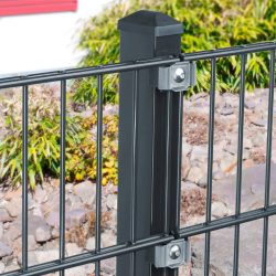 Nærbillede af en sort metal hegnspæl med to påsatte hegnspaneler, fastgjort med Klemme Beslag - Rustfrit Stål bolte. Baggrunden har et haveområde med klipper og grønne buske, sammen med en del af en rød og hvid bygning.