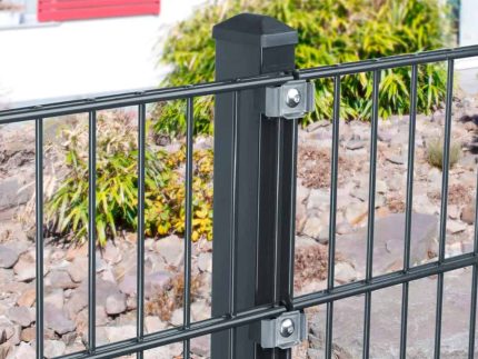 Nærbillede af en sort metal hegnspæl med to påsatte hegnspaneler, fastgjort med Klemme Beslag - Rustfrit Stål bolte. Baggrunden har et haveområde med klipper og grønne buske, sammen med en del af en rød og hvid bygning.