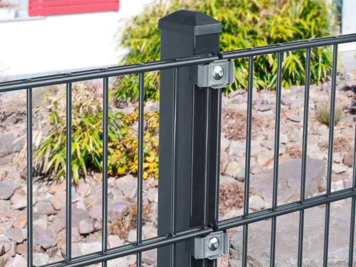 Nærbillede af en sort metal hegnspæl med to påsatte hegnspaneler, fastgjort med Klemme Beslag - Rustfrit Stål bolte. Baggrunden har et haveområde med klipper og grønne buske, sammen med en del af en rød og hvid bygning.