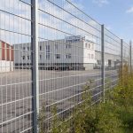 En stor, industrielt udseende bygning med flere vinduer er omkranset af et højt metalhegn lavet af den robuste Panelhegn "EXTREME" 2D 8/6/8 mm. Området inden for hegnet ser ud til at være en tom asfalteret grund, mens grøn vegetation vokser langs ydersiden. Himlen er delvist overskyet.