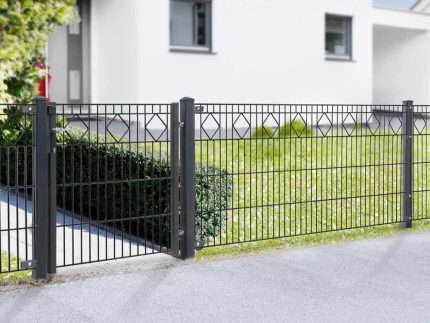 Et moderne hegn, Panelhegn med mønster Pulverlakeret Sort Metal Inkl. Stålstolper & Beslag, har lodrette stænger og et diamantformet design nær toppen. Placeret foran et hvidt, moderne hus med flere vinduer, komplementerer det den velholdte græsplæne og grønne områder, der er synlige bag hegnet.