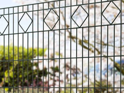 Nærbillede af en Panelhegn med mønster Pulverlakeret Sort Metal Inkl. Stålstolper & Beslag med et diamantmønster langs toppen. Bag hegnet er der slørede, blomstrende træer og grønne, grønne buske, hvilket tyder på en have eller parkomgivelse om foråret.