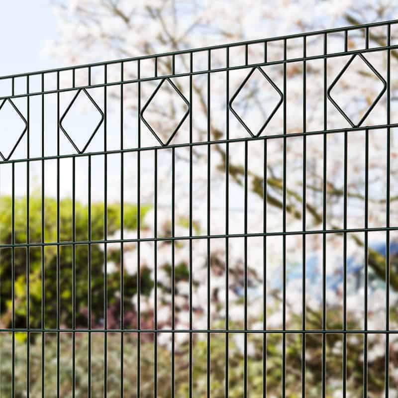 Nærbillede af en Panelhegn med mønster Pulverlakeret Sort Metal Inkl. Stålstolper & Beslag med et diamantmønster langs toppen. Bag hegnet er der slørede, blomstrende træer og grønne, grønne buske, hvilket tyder på en have eller parkomgivelse om foråret.