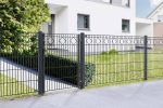 Et moderne hus med hvide vægge og grå accenter er delvist synligt i baggrunden. Forgården er omsluttet af et Panelhegn med mønster Pulverlakeret Sort Metal Inkl. Stålstolper & Beslag med geometriske diamantformer. Der er en sti, der fører fra gaden til hoveddøren, omgivet af grønt græs og buske.