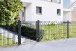 En moderne hvid bygning med store vinduer er frontet af et moderne sort metal "Panelhegn med mønster Pulverlakeret Sort Metal Inkl. Stålstolper & Beslag" hegn, med dekorative geometriske mønstre. Hegnet grænser op til en omhyggeligt vedligeholdt græsplæne med en pænt trimmet busk nær indgangsstien.