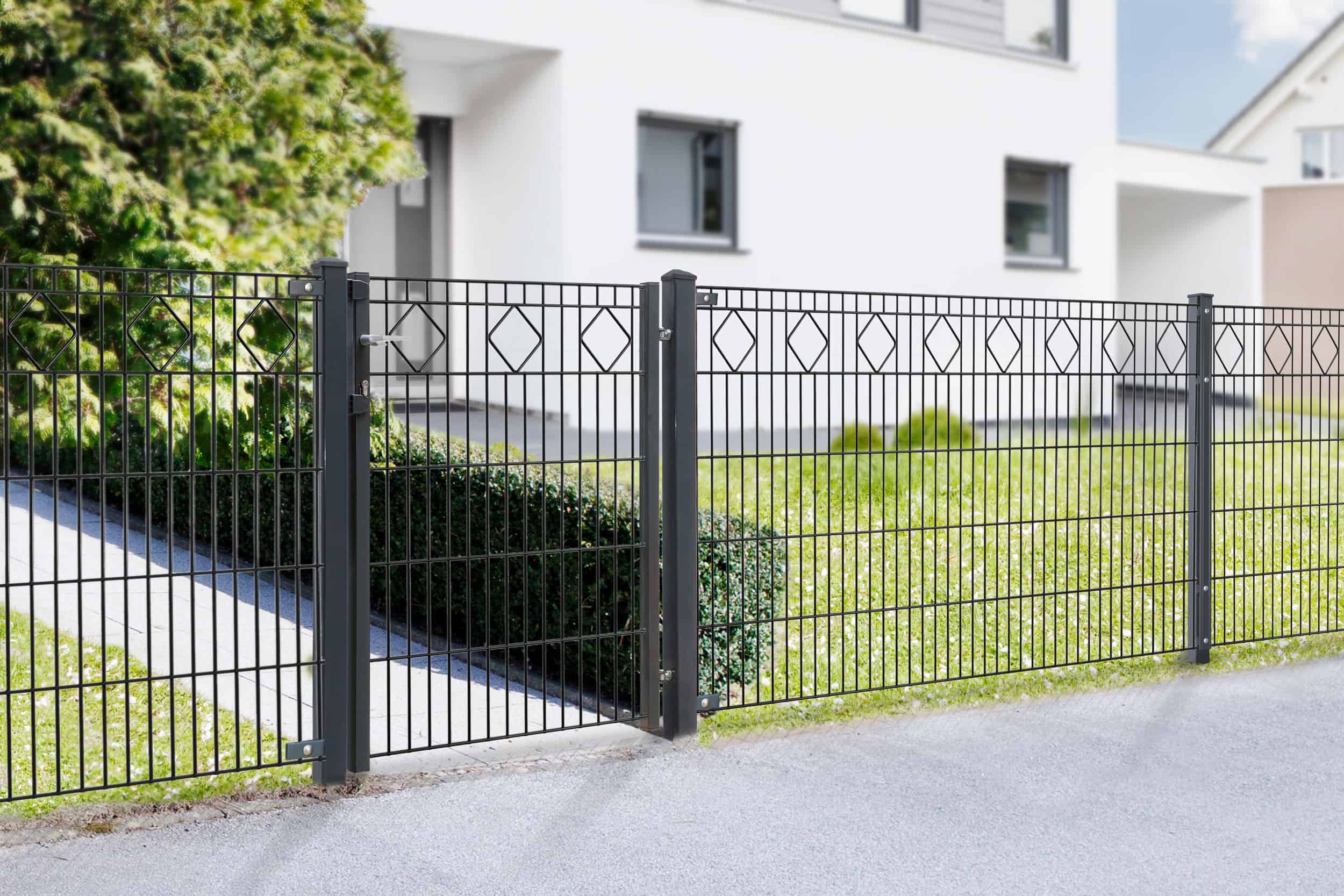 En sort **Panelhegn med mønster Pulverlakeret Sort Metal Inkl. Stålstolper & Beslag**, der viser firkantede og diamantmønstre, omslutter en pænt trimmet hæk foran et hvidt moderne hus med store vinduer. Indkørslen og fortovet er synlige sammen med en lille plet græs og et træ i baggrunden.