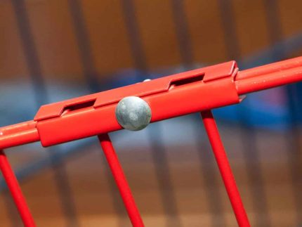 Nærbillede af et rødt metalrækværk med en vandret stang forbundet til lodrette stænger med et sølvfarvet Samlingsbeslag - Inkl. bolt og skue. Baggrunden er ude af fokus og viser en blanding af farver.