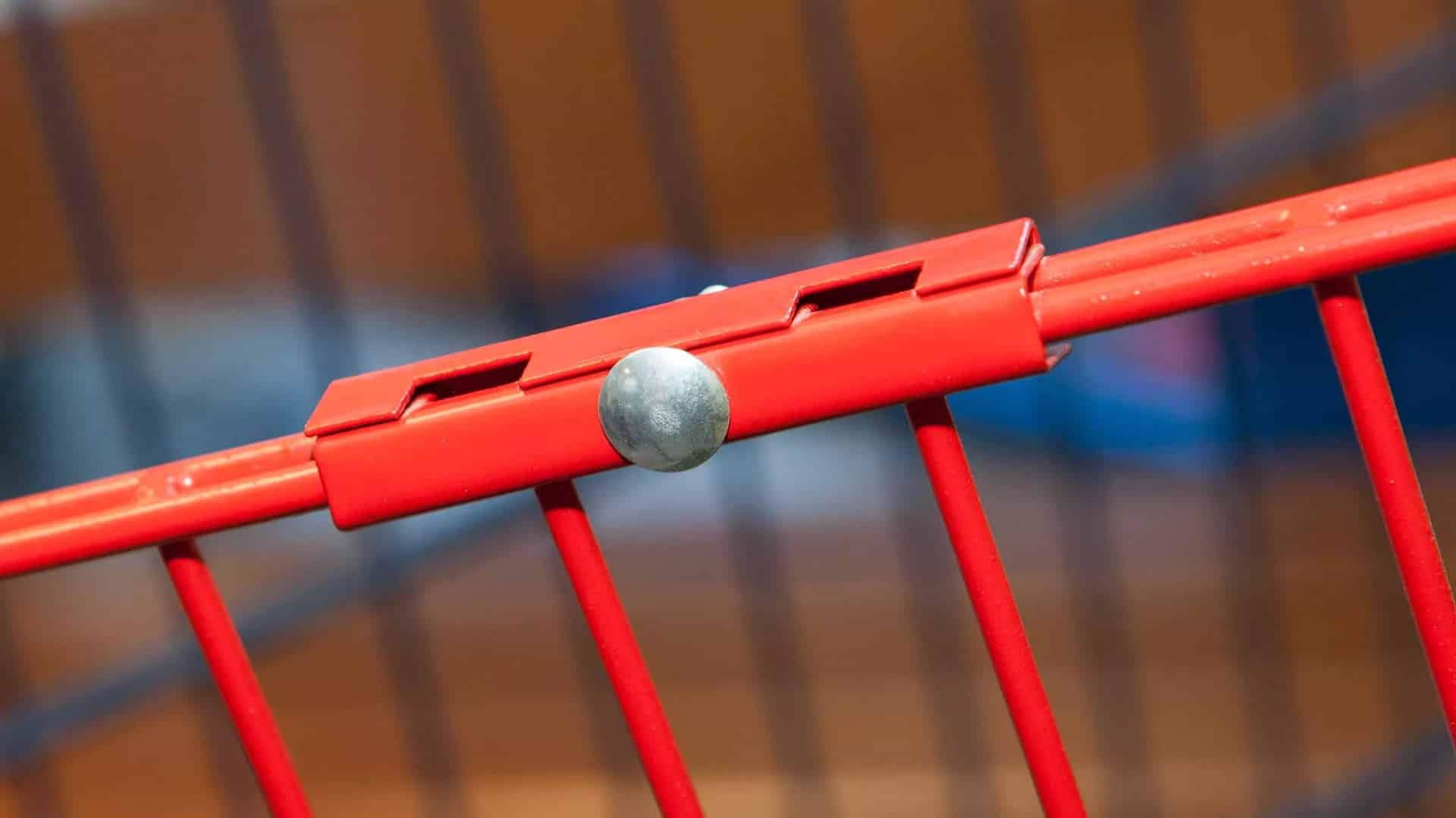 Nærbillede af et rødt metalrækværk med en vandret stang forbundet til lodrette stænger med et sølvfarvet Samlingsbeslag - Inkl. bolt og skue. Baggrunden er ude af fokus og viser en blanding af farver.