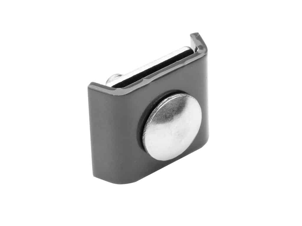 En lille, rektangulær, antrazitgrå plastikclips med en metallisk cirkulær knap i midten. Clipsen har en rille i den ene ende, formentlig til fastgørelse, og den metalliske knap rager tydeligt frem fra frontfladen.
