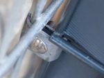 Nærbillede af en metalklemme, der fastgør to metalstænger sammen. Klemmen, der ligner en robust Hjørne Beslag - Flad, er fastgjort med en bolt og møtrik, der er synlig i midten af billedet. Stængerne og klemmen ser ud til at være en del af et strukturelt støttesystem.