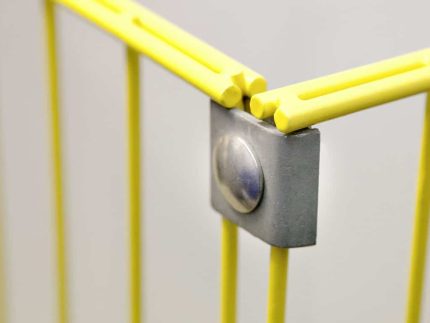 Nærbillede af hjørnet af en gul metalgenstand, der viser samlingerne forbundet med en sølvfarvet Hjørne Beslag - Flad. De gule dele inkluderer stænger og beslag, og fastgørelseselementet ser ud til at fastgøre dem i en ret vinkel. Baggrunden er sløret og grå.