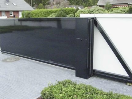 Billedet viser en Skydeport "STERK" moderne sort skydeport med lodret gitter eller lukket facade, monteret på en hvid strukturel væg. Porten, der kan variere i længden fra 3 til 20 meter, vises ved indgangen til en beboelsesejendom omgivet af grønt og placeret på en grå brolagt indkørsel. Nærliggende huse og vegetation ses i baggrunden.