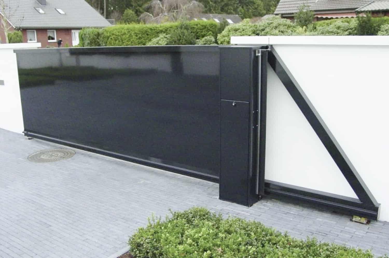 Billedet viser en Skydeport "STERK" moderne sort skydeport med lodret gitter eller lukket facade, monteret på en hvid strukturel væg. Porten, der kan variere i længden fra 3 til 20 meter, vises ved indgangen til en beboelsesejendom omgivet af grønt og placeret på en grå brolagt indkørsel. Nærliggende huse og vegetation ses i baggrunden.