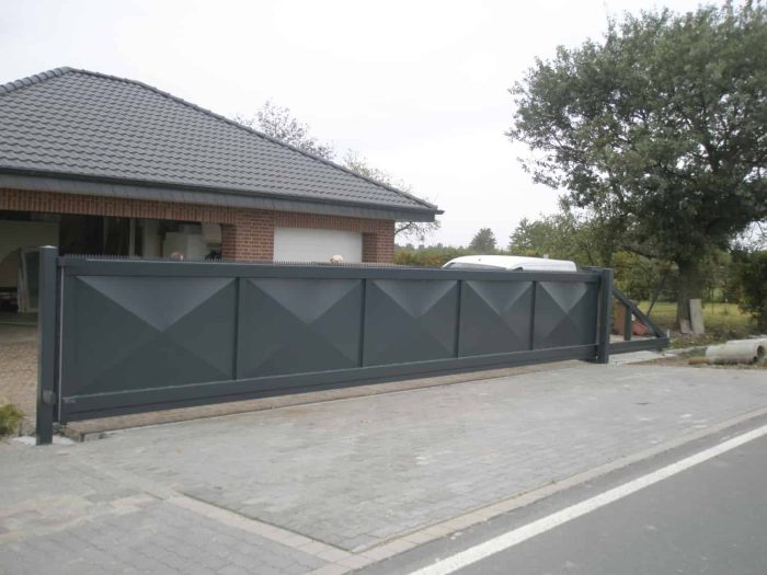 Ved indgangen til en indkørsel ved siden af et murstenshus med gråt spåntag, står en moderne Skydeport "STRONG" med en mørkegrå, lukket facade med geometriske mønstre. Den brolagte indkørsel suppleres af tilstedeværelsen af et træ og noget grønt i baggrunden, hvilket tilføjer et naturelement til scenen.