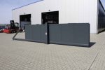 En stor industriel Skydeport "STRONG", med mørkt metal og et lodret gitter eller en lukket facade, står åben foran en moderne lagerbygning. Pakhusets lukkede facade fremviser grå vægge med flere store døre, hvoraf den ene er åben. En gaffeltruck og andet industrielt udstyr er synligt i nærheden af bygningen.