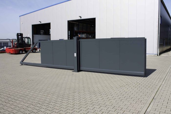 En stor industriel Skydeport "STRONG", med mørkt metal og et lodret gitter eller en lukket facade, står åben foran en moderne lagerbygning. Pakhusets lukkede facade fremviser grå vægge med flere store døre, hvoraf den ene er åben. En gaffeltruck og andet industrielt udstyr er synligt i nærheden af bygningen.