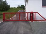En Skydeport "STRONG" med en rød finish, et lodret gitter design og en stålramme er installeret ved indgangen til en asfalteret indkørsel, der fører til en hvid bygning. Indkørslen er flankeret af græs og træer, og der er tastatur på portens stolpe til adgangskontrol.