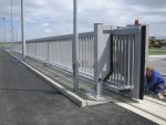En person arbejder på installationen af en Skydeport "STERK" port med en stor grå metalfacade på et industrielt eller kommercielt område. Porten, der måler mellem 3 og 20 meter, er delvist åben, og personen ser ud til at foretage justeringer eller vedligeholde mekanismen. Baggrunden viser parkerede maskiner.