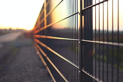 Et nærbillede af en metal Hegn taget ved solnedgang. Hegnets vandrette og lodrette linjer skaber et gittermønster, hvor solen kaster et varmt skær og lange skygger langs stængerne. Baggrunden er sløret, hvilket giver en fornemmelse af dybde og afstand, i lighed med en uddannelsesinstitutions ekspansive grund.