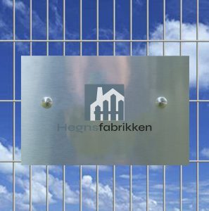 Et metalskilt monteret på et trådhegn viser et logo af stiliserede bygninger og teksten "Hegnsfabrikken", ved siden af en illustration af Samle Beslag til panelhegn - Flad, sat på baggrund af en blå himmel med spredte skyer.