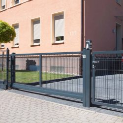 En moderne Skydeport "LIGHT" skydeport, tilgængelig i forskellige designs og længder fra 3 til 4,5 meter til både privat og industriel brug, er installeret foran en indkørsel, der fører til en garage. Metalporten med lodrette stænger står foran et ferskenfarvet hus med tre vinduer og et lille pænt beskåret træ ved siden af. Et blåt køretøj kan ses bag porten.