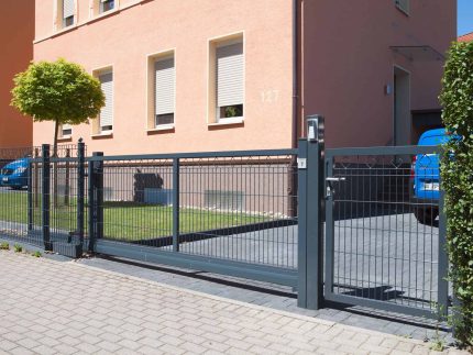 En moderne Skydeport "LIGHT" skydeport, tilgængelig i forskellige designs og længder fra 3 til 4,5 meter til både privat og industriel brug, er installeret foran en indkørsel, der fører til en garage. Metalporten med lodrette stænger står foran et ferskenfarvet hus med tre vinduer og et lille pænt beskåret træ ved siden af. Et blåt køretøj kan ses bag porten.