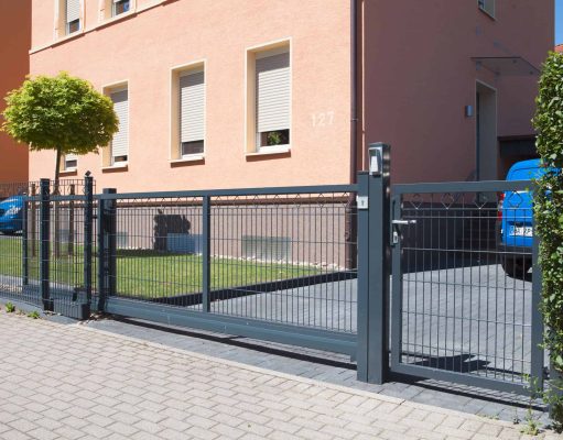 En moderne Skydeport "LIGHT" skydeport, tilgængelig i forskellige designs og længder fra 3 til 4,5 meter til både privat og industriel brug, er installeret foran en indkørsel, der fører til en garage. Metalporten med lodrette stænger står foran et ferskenfarvet hus med tre vinduer og et lille pænt beskåret træ ved siden af. Et blåt køretøj kan ses bag porten.