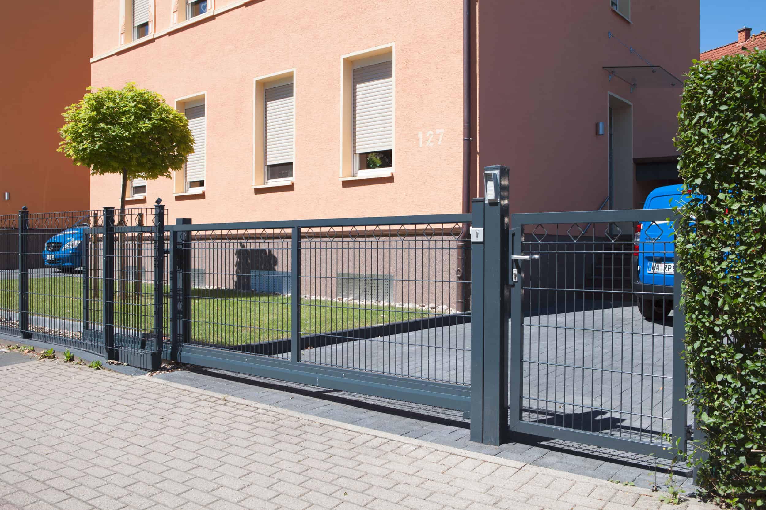 En moderne Skydeport "LIGHT" skydeport, tilgængelig i forskellige designs og længder fra 3 til 4,5 meter til både privat og industriel brug, er installeret foran en indkørsel, der fører til en garage. Metalporten med lodrette stænger står foran et ferskenfarvet hus med tre vinduer og et lille pænt beskåret træ ved siden af. Et blåt køretøj kan ses bag porten.