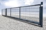 En lang, mørk metal Skydeport "LIGHT" skydeport, fås i en længde på 3 til 4,5 meter og velegnet til både privat og industriel brug, står lukket på en grus overflade. Porten har et gitterlignende mønster af lodrette og vandrette stænger, understøttet af en robust ramme og en enkelt stolpe. Baggrunden viser en overskyet himmel, hvilket skaber en skarp kontrast til portens industrielle design.