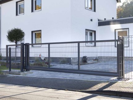Et moderne hus med hvide vægge har en stor indkørsel sikret af en robust, grå metal Skydeport "V-Star EXTREME" 2D Panelfyld 8/6/8 MM. Gården er pænt anlagt med et lille træ og forskellige planter. Baggrunden viser en del af en tilstødende sort bygning og noget grønt.