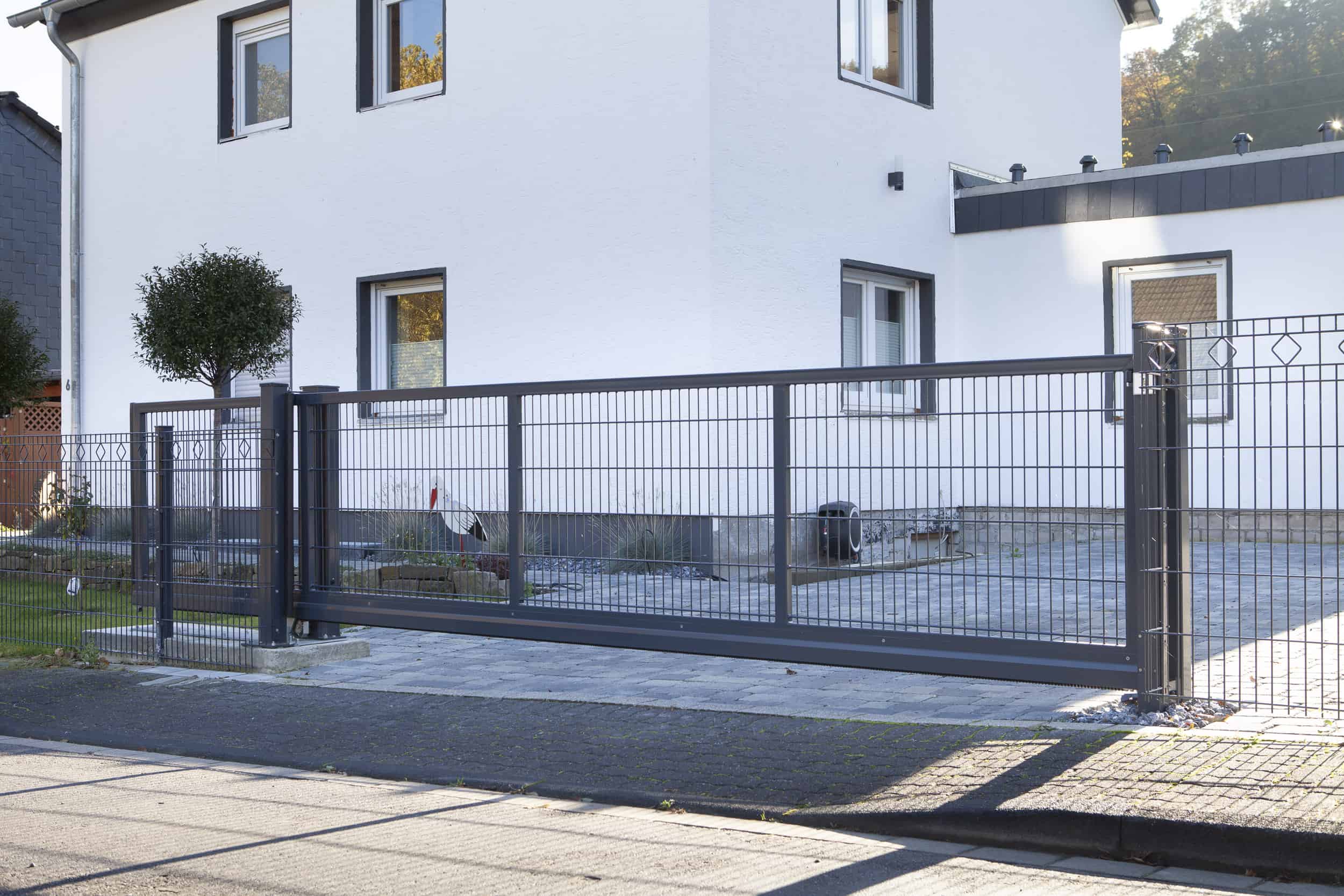 Et moderne hus med hvide vægge har en stor indkørsel sikret af en robust, grå metal Skydeport "V-Star EXTREME" 2D Panelfyld 8/6/8 MM. Gården er pænt anlagt med et lille træ og forskellige planter. Baggrunden viser en del af en tilstødende sort bygning og noget grønt.