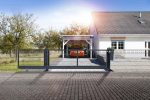 Et moderne hvidt enplanshus med tegltag ses med en bil parkeret under en carport. Huset er omgivet af et metalhegn med Skydeport "V-Star EXTREME" 2D Panelfyld 8/6/8 MM og en Skydeport skydeport Solen skinner og kaster skarpt lys over scenen, og træer er i baggrunden.