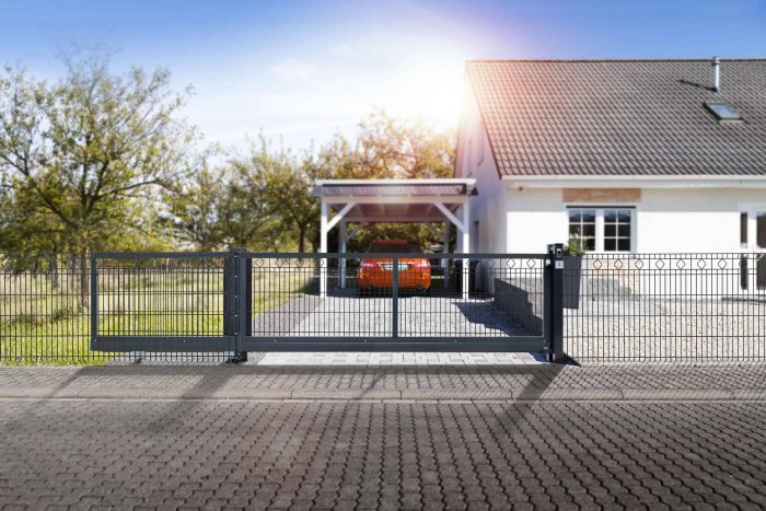 Et moderne hvidt enplanshus med tegltag ses med en bil parkeret under en carport. Huset er omgivet af et metalhegn med Skydeport "V-Star EXTREME" 2D Panelfyld 8/6/8 MM og en Skydeport skydeport Solen skinner og kaster skarpt lys over scenen, og træer er i baggrunden.
