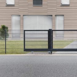 Et moderne hus med stor Skydeport "V-Star EXTREME" 2D Panelfyld 8/6/8 MM metalskydeport foran Huset har store vinduespartier med lukkede persienner og en velholdt græsplæne med diverse buske og træer. Til højre , en tilstødende bygning med orange accenter er delvist synlig.