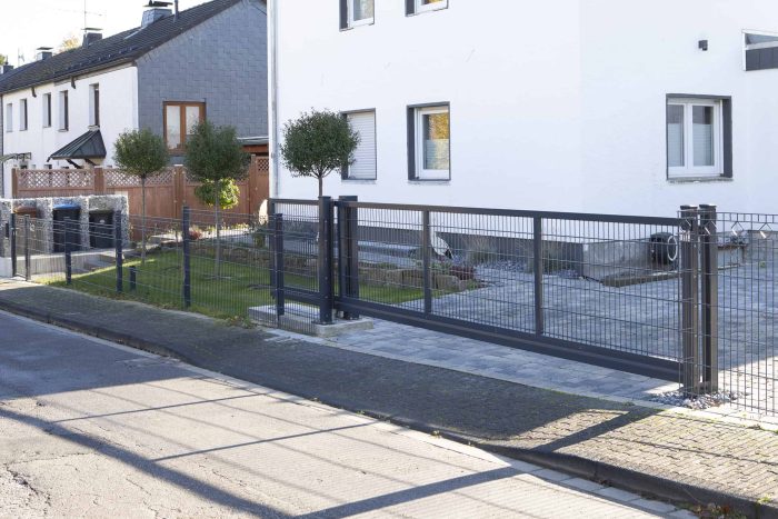 En moderne boliggade med et hvidt to-etagers hus med gråt tag og accenter af Skydeport "V-Star EXTREME" 2D Panelfyld 8/6/8 MM. Forhaven har en minimalistisk have med et ensomt træ, sikret af et sort metalhegn med skydeport. Nabohuse viser lignende moderne arkitektur.